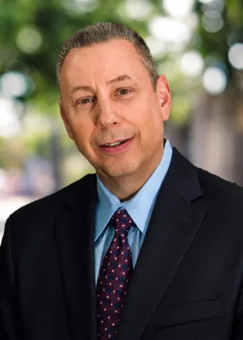Richard wears a dark suit, blue shirt, and patterned tie in a head-and-shoulders portrait.