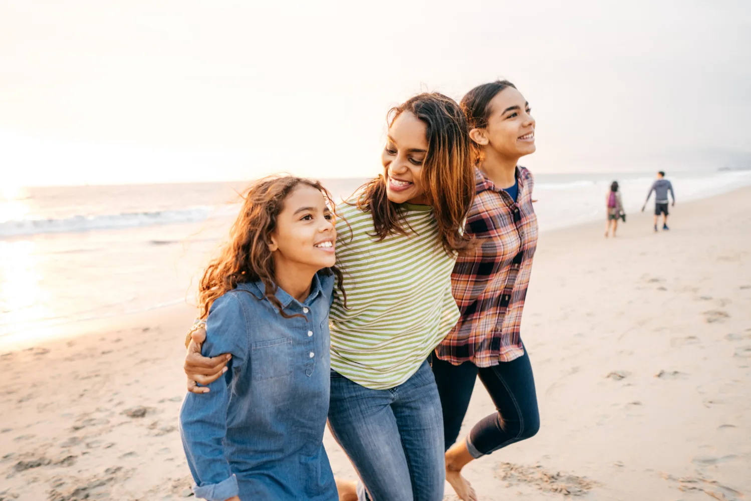 A mother embraces her two daughters while walking along a beach at sunset.