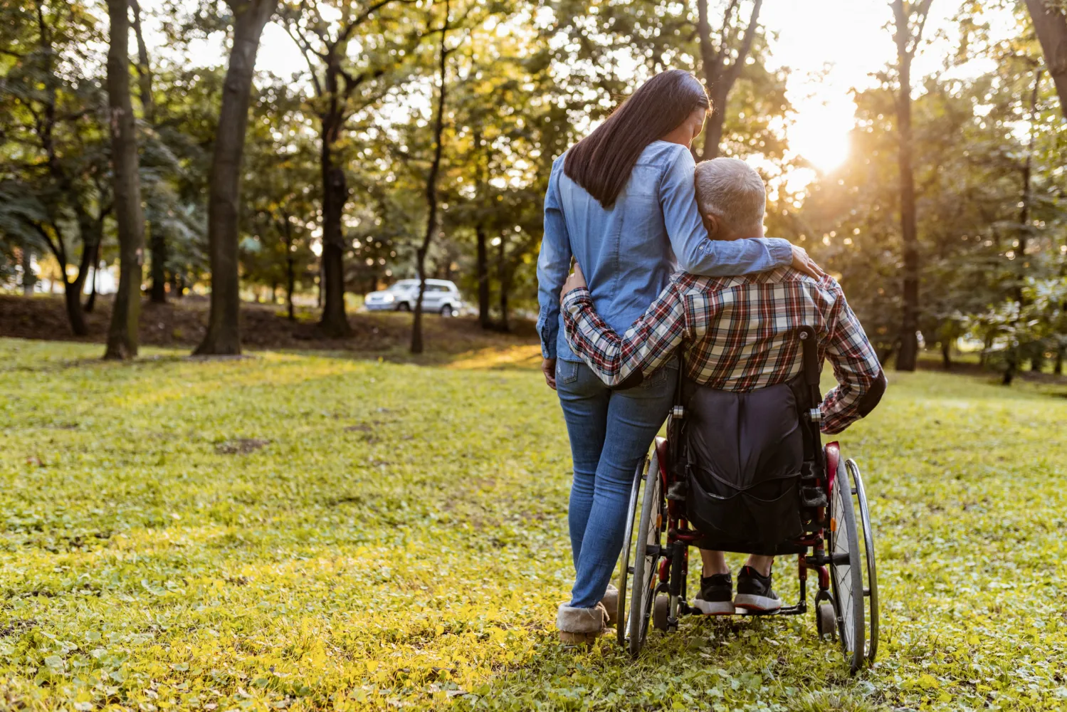 Woman standing beside and embracing an older man in a wheelchair as they enjoy a sunlit park.