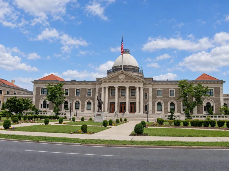 Neoclassical courthouse with a central dome, U.S. flag, front columns, statue, and landscaped gardens beside a roadway.