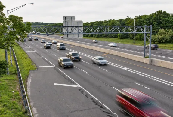 Multiple cars travel along a divided multi-lane highway beneath an overhead sign gantry, with trees lining both sides.